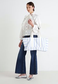 Mulher vestindo uma blusa branca com babados, jeans de pernas largas, sapatilhas prateadas e segurando uma grande bolsa listrada em azul e branco.