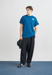 Μπλε βαμβακερό T-shirt με κοντά μανίκια και μικρό λογότυπο στο στήθος, σε συνδυασμό με χαλαρές μαύρες παντελόνες και μαύρα αθλητικά παπούτσια με λευκές λεπτομέρειες.