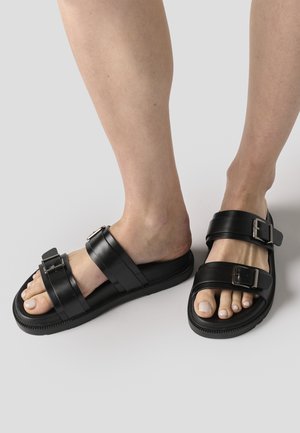 Pies con sandalias negras tipo slide con dos correas ajustables con hebillas sobre un fondo claro.