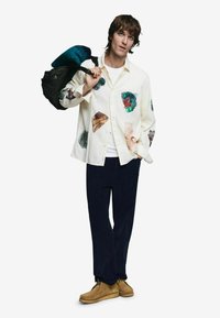 Valge nööbitav särk, millel on värvikirevad kestade ja loodusteemalised plaastrid, kantud tumedate pükstega ja beežide kingadega, ning kaasas musta ja türkiissinise kotiga.