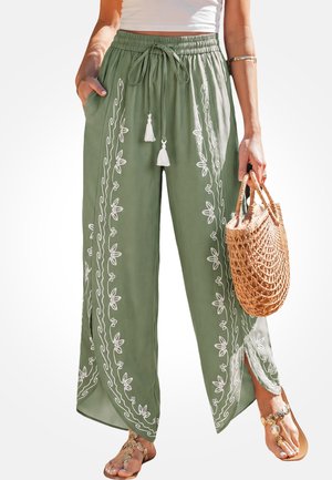 Vrouw draagt olijfgroene wijde broek met witte bloemenborduursel, houdt een gevlochten handtas vast en draagt sandalen met decoratieve riempjes.