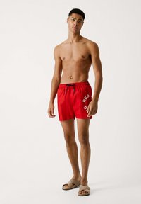 Bañador rojo con cintura elástica y cordón. Presenta un logo en blanco contrastante en el muslo izquierdo. Textura suave, diseño hasta la rodilla.