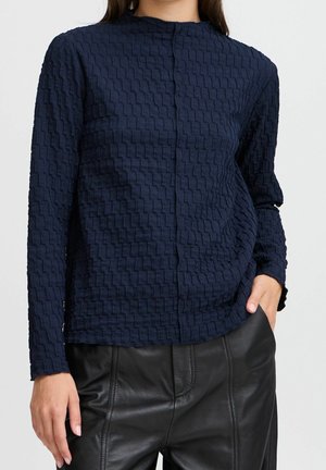 Woman wearing a navy textured long-sleeve top and black leather pants with one hand in pocket against a plain light background.