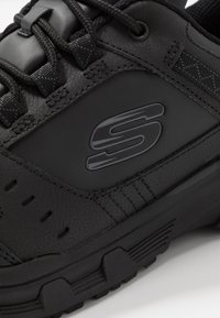 Black athletic shoe featuring a textured synthetic upper, logo detail, sturdy laces, and a rugged rubber sole with traction grooves.