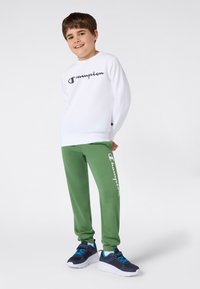 Smilende gutt står, iført hvit hettegenser og grønne joggebukser med "Champion" merking, samt marineblå joggesko med blå snøring.