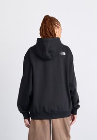 Personne aux cheveux tressés portant un sweat à capuche noir The North Face et un pantalon marron, vue de dos sur un fond uni.