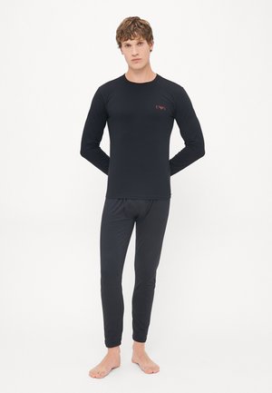 Schwarzes, langärmliges Thermooberteil und taillierte Hose aus glattem Stoff. Mit einem kleinen roten Logo auf der Brust. Barfußmodell steht mit den Händen hinten.