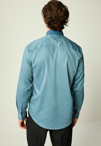 Cămașă turquoise cu mânecă lungă, dintr-un material texturat și cu un guler patternat. Prezintă manșete cu nasturi și un tiv curbat.