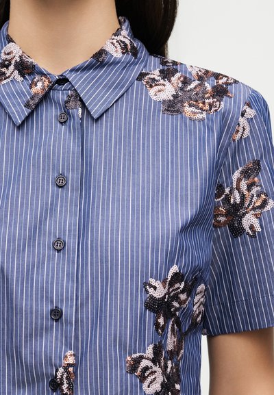 Chemise bleue à manches courtes boutonnée avec fines rayures blanches et motifs floraux à sequins noirs, blancs et marron sur le col et les manches.