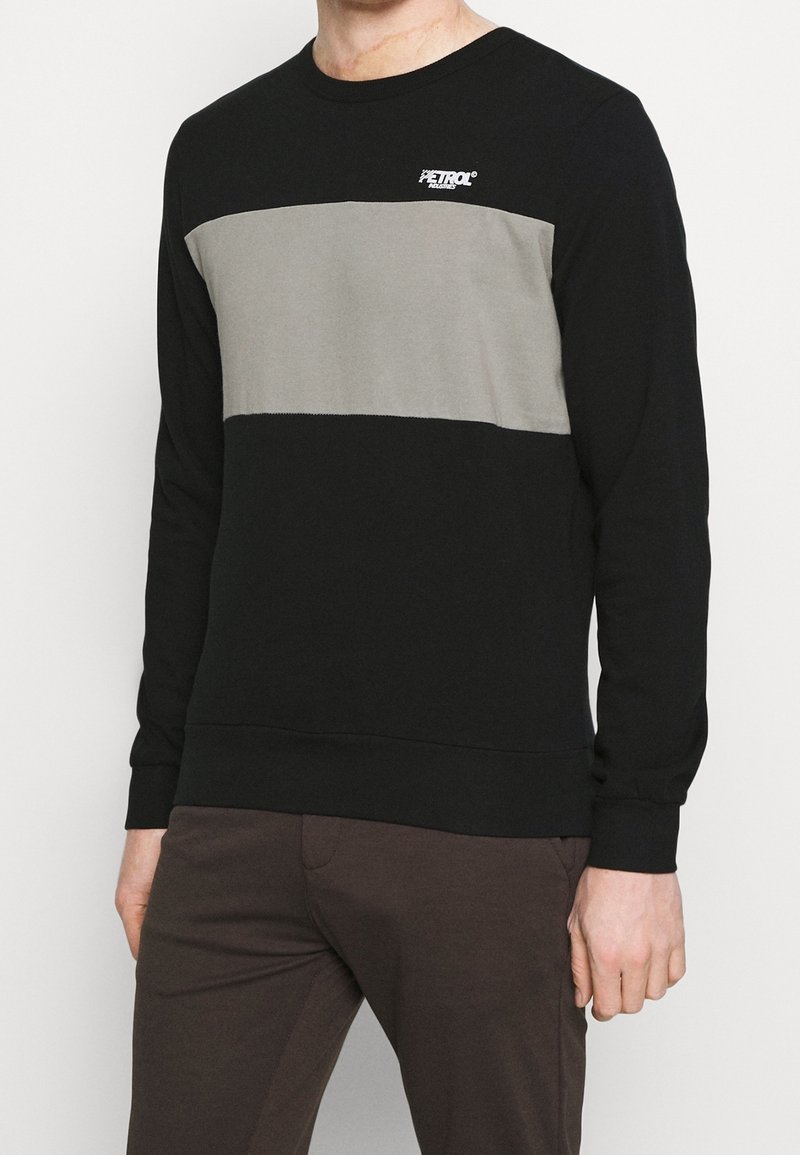 Homme portant un sweat-shirt noir et gris à manches longues avec le logo "Petrol" sur la poitrine et un pantalon marron foncé, debout devant un fond blanc.