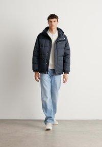 Νεαρός άνδρας που περπατά σε εσωτερικό χώρο, φορώντας μαύρο μπουφάν, λευκό πόλο, ανοιχτό μπλε τζιν και λευκά παπούτσια, μπροστά σε έναν απλό λευκό τοίχο.