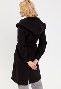 Manteau noir mi-long en mélange de laine, avec capuche, ceinture et manches longues. Texture lisse avec un design épuré.