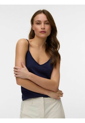 Žena s dlhými hnedými vlasmi oblečená v námorníckom modrom tielku a béžových nohaviciach, so založenými rukami a hľadí do kamery na jednofarebnom pozadí.