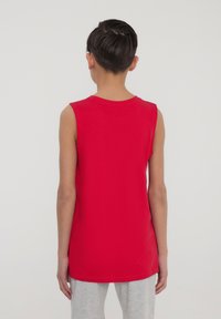 Maglietta rossa senza maniche in cotone, con scollatura rotonda e texture liscia, indossata con pantaloni tuta grigi.