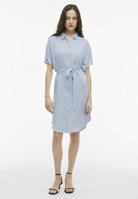 Vestito camicia azzurro chiaro con maniche corte, chiusura con bottoni anteriore, vita legata e orlo curvo, realizzato in tessuto liscio. Accentuato con sandali.