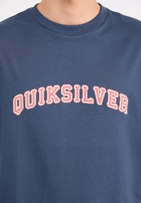 Κοντινό πλάνο ενός ατόμου που φοράει ένα ναυτικό μπλε T-shirt με τη λέξη "QUIKSILVER" σε έντονους, ανοιχτό ροζ περιγραμμένους χαρακτήρες επάνω στο στήθος.