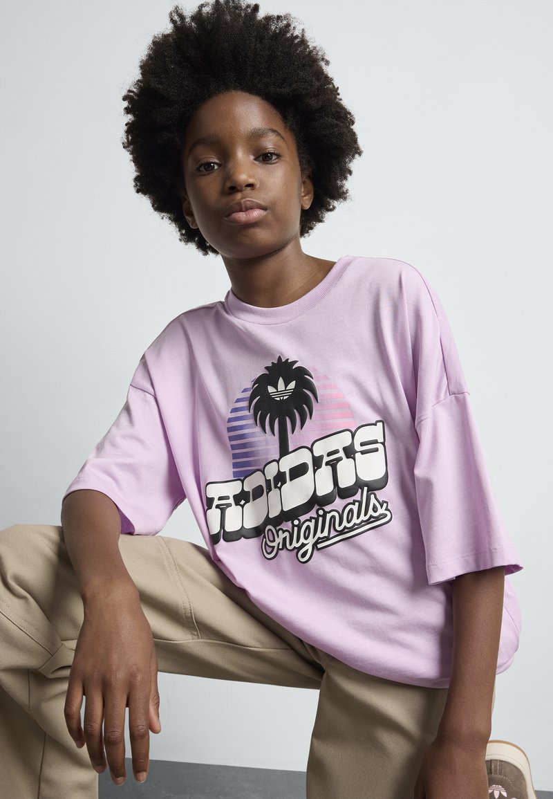 Jaunas žmogus su natūraliais plaukais vilki per didelę rožinę "Adidas Originals" marškinėlį ir bežemius kelnes, sėdi su vienu keliu pakeltu prieš paprastą foną.