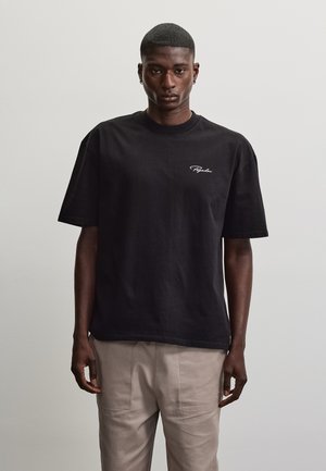 Mann iført svart oversized t-skjorte med lite hvitt logo på brystet og beige bukser, står foran ensfarget grå bakgrunn.
