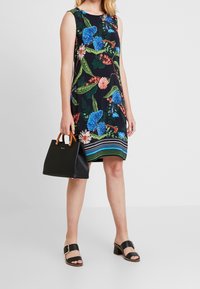 Femme portant une robe sans manches noire à fleurs, tenant un sac à main noir avec des anses marron, chauss ée de sandales à talons noires avec des brides à boucle.