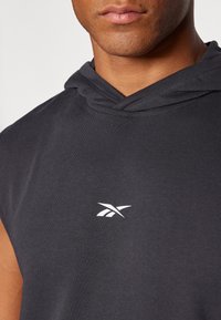 Hombre con un hoodie negro sin mangas y un pequeño logo blanco de Reebok en el pecho, recortado debajo de la nariz.