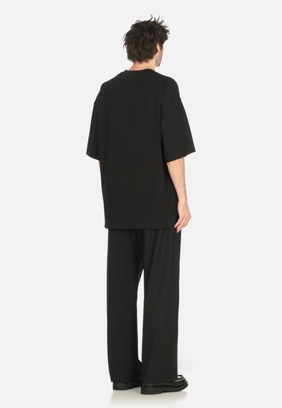 T-shirt nera oversize con maniche corte, realizzata in tessuto morbido. Indossata con pantaloni larghi neri coordinati e scarpe basse, vista da dietro.