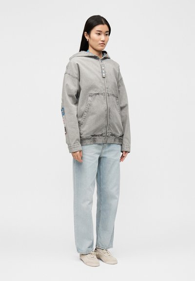 Γυναίκα στέκεται φορώντας γκρι φούτερ με φερμουάρ και μπαλώματα στο μανίκι, ανοιχτό μπλε φαρδιά τζιν και μπεζ αθλητικά παπούτσια μπροστά σε απλή φόντο.