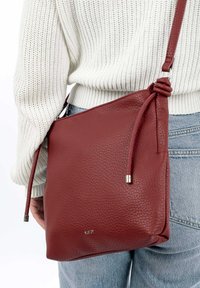 Sac bandoulière en cuir rouge avec une surface texturée, doté d'une sangle nouée et d'accents en métal argenté. Forme rectangulaire compacte.