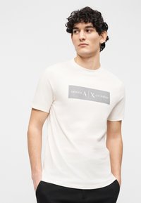 Νεαρός άνδρας με σγουρά μαλλιά, που φοράει λευκό μπλουζάκι Armani Exchange και μαύρο παντελόνι, κοιτάζοντας πλευρικά με τα χέρια στις τσέπες σε γκρι φόντο.