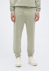 Άνδρας που φοράει ανοιχτοπράσινο παντελόνι φόρμας με κορδόνι και λευκά αθλητικά παπούτσια, στέκεται μπροστά σε απλό λευκό φόντο.