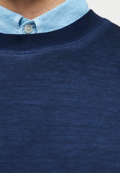 Μπλε ναυτικό πουλόβερ με λαιμόκοψη, με ομαλή υφή, που συνδυάζεται με ένα γαλάζιο πουκάμισο με γιακά από κάτω και ένα κουμπί στον γιακά.