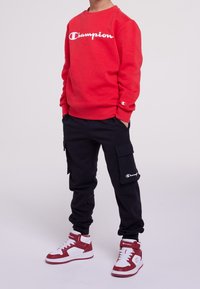 Felpa rossa con logo "Champion" bianco, pantaloni cargo neri con tasche laterali e sneaker alte bianche e rosse.