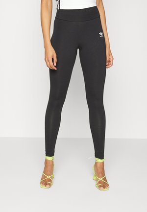 Leggings nere con vita alta, design aderente e tessuto testurizzato. Presentano un piccolo logo bianco. Tacchi gialli luminosi con cinturini.
