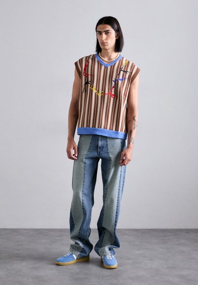 Top senza maniche a righe marroni con dettagli ricamati colorati, abbinato a jeans in denim splittato azzurri e verdi e scarpe da ginnastica blu.