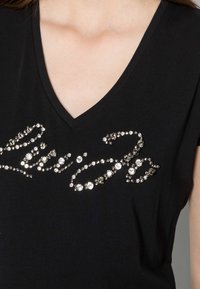 Camiseta negra de cuello en V con diseño cursivo de strass metálicos, con tamaños y tonalidades variables para crear contraste con la tela.