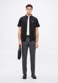 Μαύρο πουκάμισο με κοντά μανίκια και λογότυπο, λευκό T-shirt από μέσα, γκρι παντελόνι κανονικής ραφής, μαύρα δερμάτινα παπούτσια και μαύρη τσάντα χειρός.