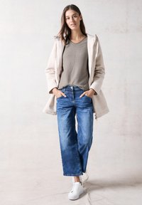 Giacca in pile beige chiaro, top grigio a coste, jeans blu larghi e sneakers bianche. Tessuto strutturato, vestibilità comoda con cappuccio e tasche frontali.