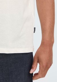 Camiseta de algodón color crema con una abertura lateral y una pequeña etiqueta negra, combinada con jeans de mezclilla oscuros. Textura suave y corte clásico.