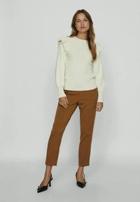 Sweater color creme com detalhes em babados nos ombros, combinado com calças justas castanhas e sapatos de salto alto pretos.