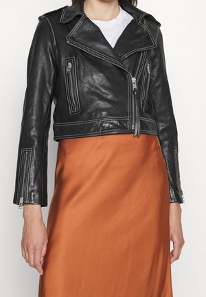 Femme portant une veste en cuir noire courte avec des fermetures éclair argentées sur un top blanc et une jupe orange.