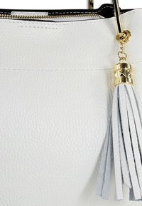 Sac à main en cuir blanc avec une surface texturée, des éléments métalliques dorés et un pompon en cuir gris attaché à la fermeture éclair.