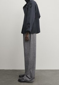 Granatowa kurtka z dużymi guzikami i wysokim kołnierzem, zestawiona z szerokimi szarymi spodniami. Model nosi czarne buty, stoi na neutralnym tle.