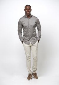 Grijze langemouwen shirt met knopen, lichtbeige broek en bruine sneakers. Model staat met handen in de zakken tegen een witte achtergrond.