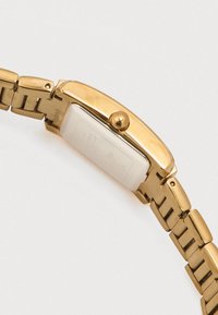 Horloge met gouden tint, rechthoekige wijzerplaat, glad metalen bandje en een kroon aan de zijkant. De achterkant toont gegraveerde tekst en logogegevens.