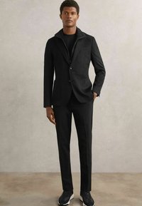 Sort skræddersyet jakke med høj krave og enkelt knap, parret med tætsiddende bukser og sorte sneakers. Glat stof, moderne design.