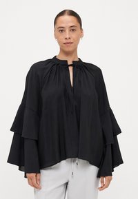 Black blouse with layered, bell-shaped sleeves. Features a round neckline with a tie, crafted from a smooth, lightweight fabric.