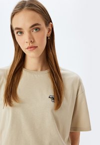 Beige bomulds T-shirt med en koala grafik broderet på brystet. Korte ærmer og klassisk rund halsudskæring. Blød tekstur.