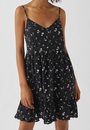 Femme portant une robe noire sans manches à fines bretelles avec un petit imprimé floral blanc et violet, debout devant un fond uni.