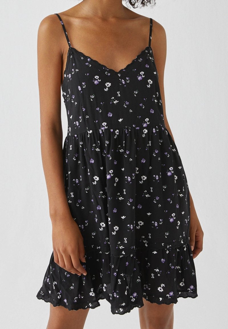 Femme portant une robe noire sans manches à fines bretelles avec un petit imprimé floral blanc et violet, debout devant un fond uni.