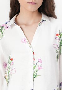 White button-up shirt featuring a floral pattern with multicolored flowers. Made of lightweight fabric, it has a classic collar and soft texture.