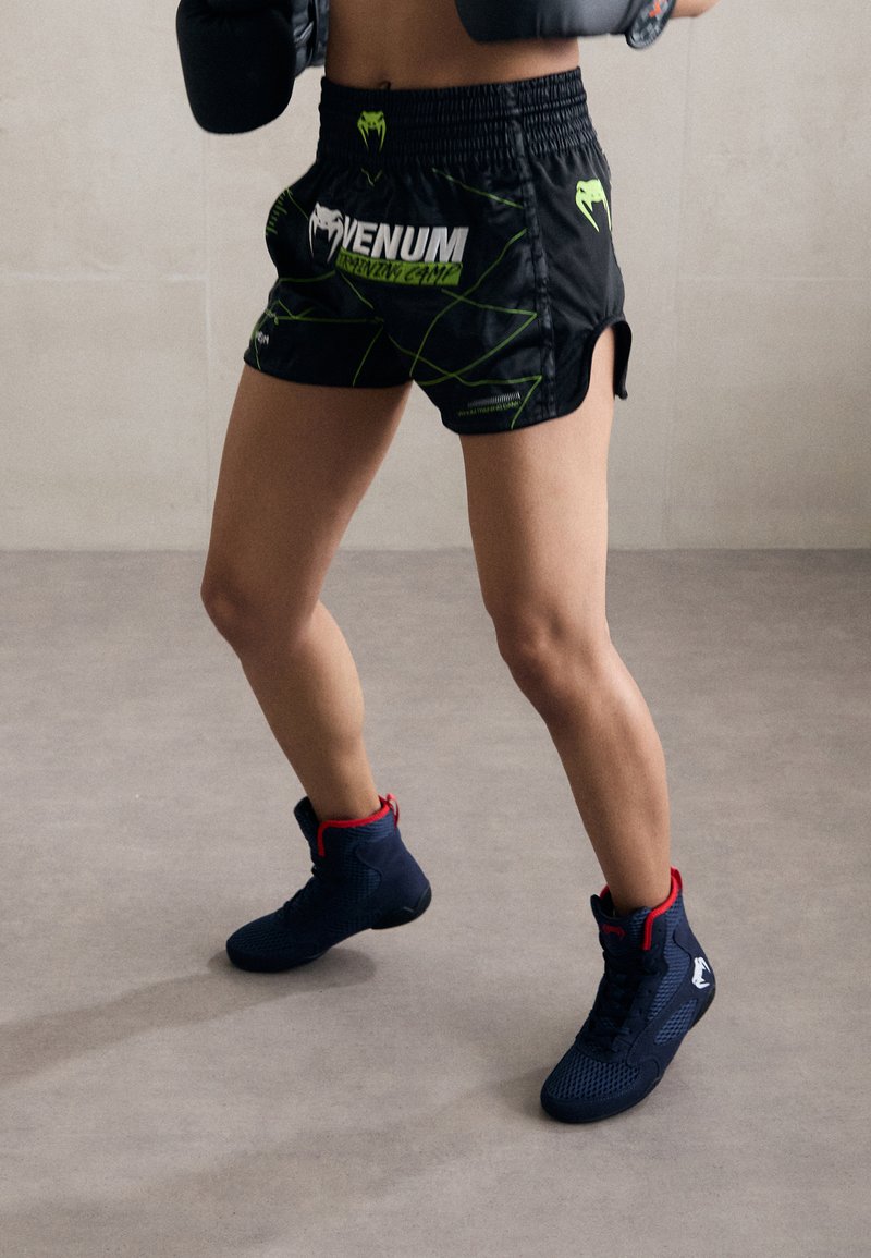 Black athletic shorts with green accents and logo, paired with navy footwear featuring red details. Focus on performance and movement.